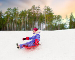 childhood, sledging and season concept - happy little girl sliding down on snow saucer sled outdoors in winter over snowy park or forest background
