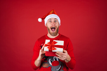 Red-bearded man in a santa hat and a christmas sweater on a red background with a gift box is excited, looking forward to the holiday