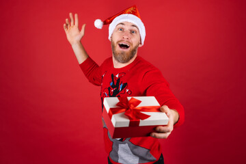 Red-bearded man in a santa hat and a christmas sweater on a red background with a gift box is excited, looking forward to the holiday