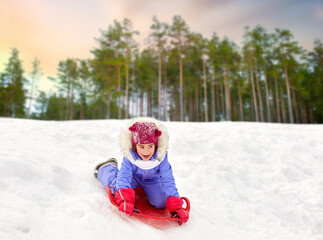 childhood, sledging and season concept - happy little girl sliding down hill on snow saucer sled outdoors in winter over snowy forest or park background