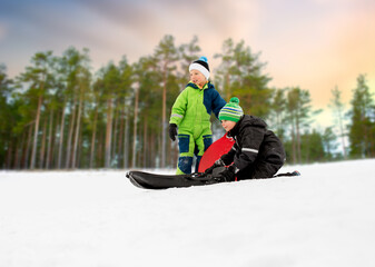 Fototapeta na wymiar childhood, sledging and season concept - little boys sliding on sleds down snow hill in winter over snowy forest or park background