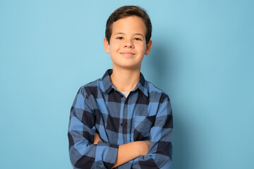 Smiling boy wearing plaid shirt with arms folded standing over blue background.