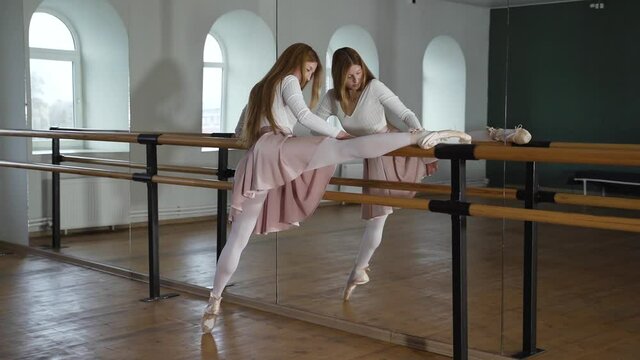 Young brunette woman in white tights is doing stretching in the dance hall near the mirror. Ballerina practicing splits