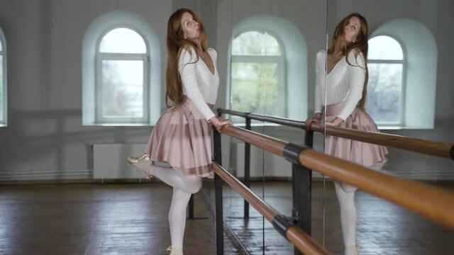 Ballerina in pointe shoes dances near mirror in dance studio. Young woman in pink skirt and white tights is dancing