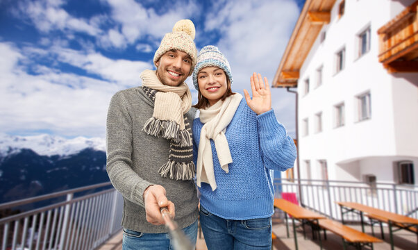 travel, tourism and winter holidays concept - happy couple in knitted hats and scarves taking picture by selfie stick over mountains and ski resort background