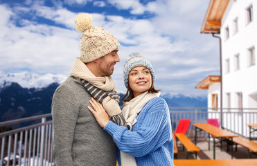 travel, tourism and winter holidays concept - happy couple in knitted hats and scarves over mountains and ski resort background