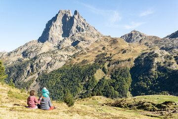 Family day at the mountains in  french Pyrenees. Father, mother and baby sitting on a meadow while enjoy views of Pic du Midi d'Ossau. Mountain travel vacation. - obrazy, fototapety, plakaty