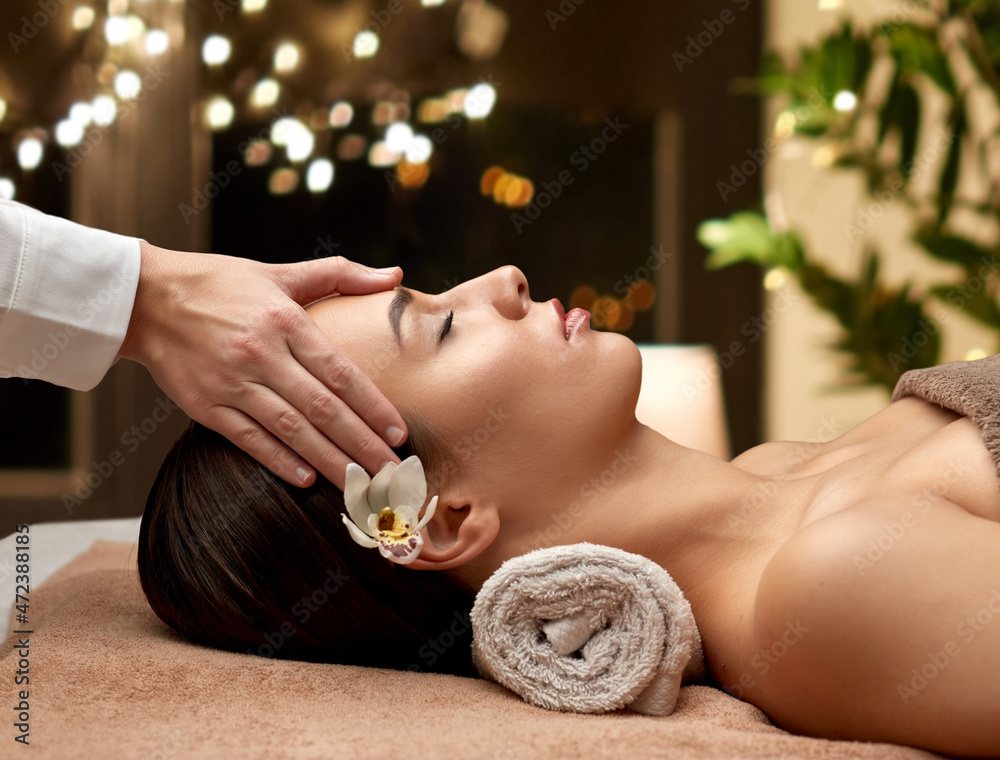 Wall mural wellness, beauty and relaxation concept - beautiful young woman lying with closed eyes and having face and head massage at spa over christmas lights on window background