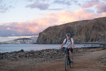 Happy senior cyclist man at the beach at sunset enjoying freedom and beauty in nature. Elderly man with protective helmet and backpack