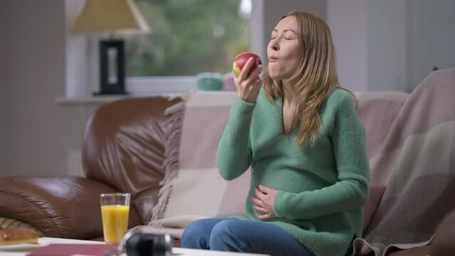 Medium shot smiling pregnant young woman eating apple smiling stroking belly. Portrait of Caucasian expectant enjoying taste of healthful fruit sitting on comfortable couch in living room. Slow motion