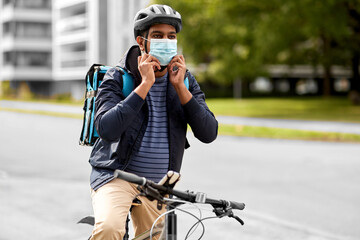 food shipping, health and people concept - indian delivery man in bike helmet and mask with thermal insulated bag riding bicycle on city street