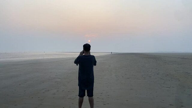 Rear cinematic shot of a man standing and clicking picture of the sunset at a lonely beach.