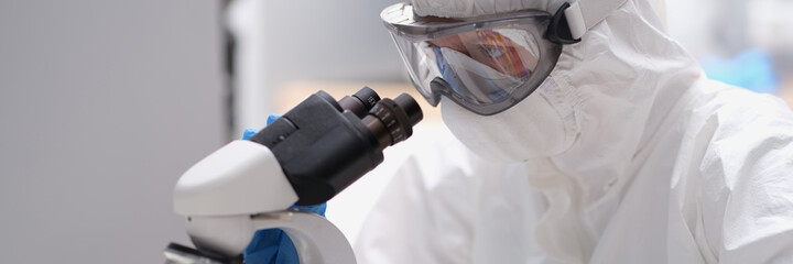 Scientist in protective suit and mask looks through microscope
