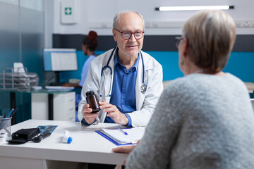 Fototapeta premium Doctor giving bottle of pills with medicine to old patient at meeting appointment in cabinet. Physician showing jar with prescription treatment to sick woman at medical checkup visit.