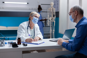 Woman doctor with face mask taking notes on papers at medical consultation with man. Physician signing clipboard files for healthcare treatment and medicine during covid 19 pandemic