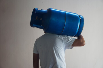 Back view of man carries blue LPG gas cylinder tank on his shoulder to make delivery to local...
