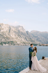 Groom hugs bride on the pier against the background of mountains