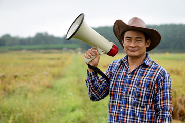 Asian male farmer hold megaphone, stands at paddy field. Concept : farmer want to communicate something to government to help and support agricultural crops. Protest. Protester. Announce to the world.