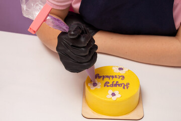 Hand in glove holds pastry bag with cream, the inscription Happy Birthday is applied to bento cake. Selective focus.