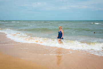 Happy mature woman enjoying on the sea beach, walking on the waves. An elderly woman in a dark blue swimsuit on the seashore.