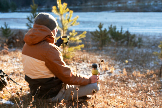 Young Man Drinking Tea From Thermos By River Covered With First Snow Ice On Hike In Fall