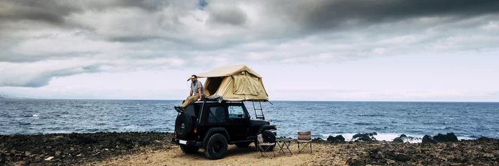 Foto op Plexiglas Banner reiskop met man zittend op het dak van de auto met kampeertent die tijd heeft om te ontspannen en te genieten van een avontuurlijke reislevensstijl of alternatieve gratis vakantie © simona
