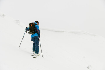 Fototapeta na wymiar rear view of man skier with trekking poles on snowy hill with cloudy sky in the background.