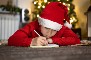 Cute boy in red sweater and red santa hat writes a letter to Santa and thinking about presents