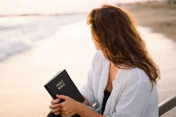 Christian woman holds bible in her hands. Reading the Holy Bible on the sea during beautiful sunset. Concept for faith, spirituality and religion. Peace, hope