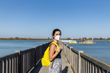 young woman traveler with yellow backpack and face mask on a pier on her way to her next trip