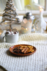 Fototapeta na wymiar Plate with cinnamon, anise and dry oranges, cozy blanket, various Christmas deocrations and lit candles. Hygge at home. Selective focus.