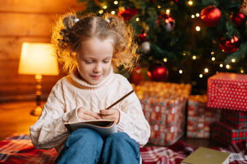 Fototapeta na wymiar Close-up of adorable dreaming little blonde curly child girl writing letter to Santa Claus sitting on floor on background of Christmas tree and gift boxes, in room with festive red bright interior