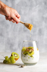 Plain yoghurt with pieces of fresh kiwi fruits, white grapes, granola, mint leaves in a glass, and...