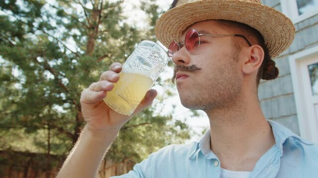 Old fashioned fancy young hipster man with moustache in straw hat, pink sunglasses drink fresh pineapple juicein villgae summer country house. Morining of modern outdoor freelance lifestyle
