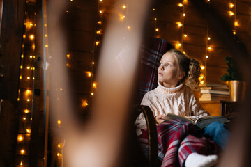 Pretty little blonde curly child girl wrapped in plaid looking away holding book sitting in rocking chair at dark room with festive interior, background of wall decorated Christmas bokeh lights. © dikushin