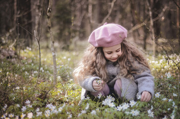 Picture of a girl in a beret and coat sitting on her haunches in the grass and flowers
