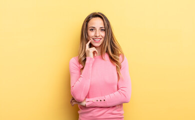 pretty caucasian woman looking happy and smiling with hand on chin, wondering or asking a question,...