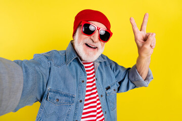 Self-portrait of elderly retired pensioner cheerful grey-haired man showing v-sign isolated over bright yellow color background