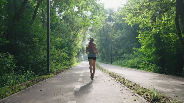 Female jogger wearing top and shorts running on alley in park in slow motion, sun shining through green foliage. Following shot fit woman exercising outside. Concept of healthy lifestyle