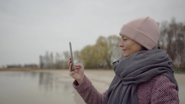 middle aged woman stands by the sea in winter and takes pictures of the landscape with her cell phone
