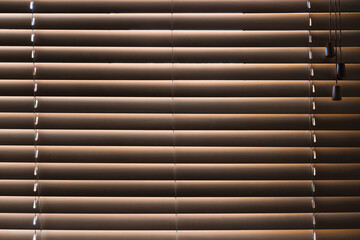 Obraz premium A look from a dark room at a window closed by brown blinds. Light from the street shines through the closed wooden slats. Background. Texture.