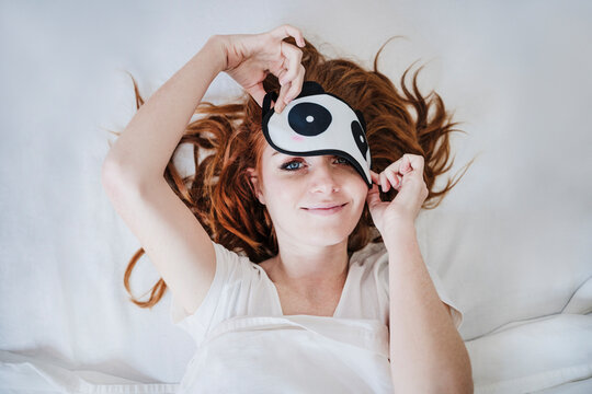 Smiling woman removing eye mask while lying on bed at home