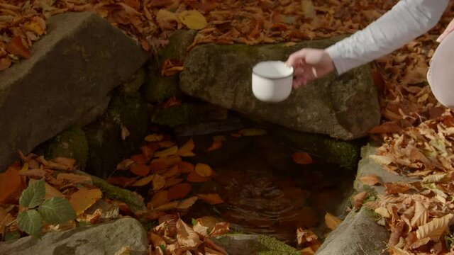 Close-up of womans hand filling a mug with water from a water source. Fallen leaves on the background.