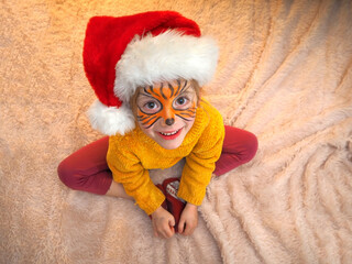 A cheerful child in a tiger mask and Santa Claus hat sits on the bed. New year of the tiger. New Year celebration. Carnival. - Powered by Adobe