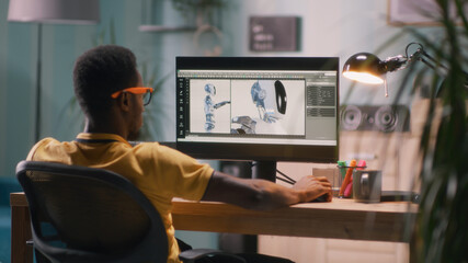 Black man in glasses and yellow t shirt sitting at table and using computer to create 3D model of futuristic robot for movie during work in home office