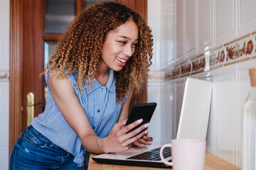 Smiling curly haired woman with mobile phone using laptop at home