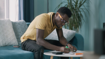 African American guy in glasses calculating utility bills and taxes and getting upset while sitting...