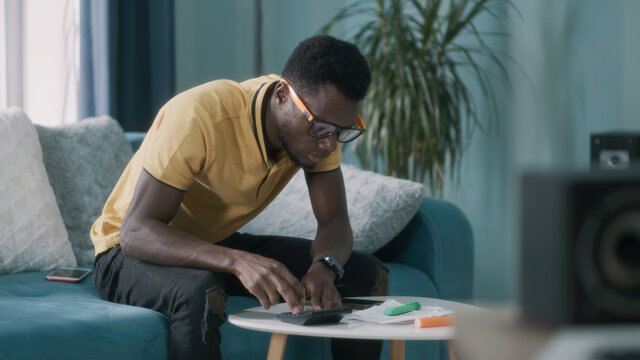 African American guy in glasses calculating utility bills and taxes and getting upset while sitting on sofa at home