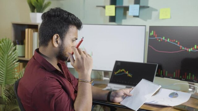 Investor thinking or analyzing stock market by writing notes in front of live share market charts on monitor- Concept of planning,making strategy and investing money on equity shares