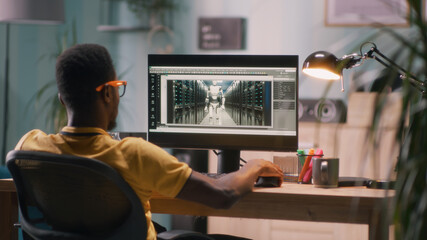 Black man in glasses and yellow t shirt sitting at table and using computer to create 3D model of futuristic robot for movie during work in home office